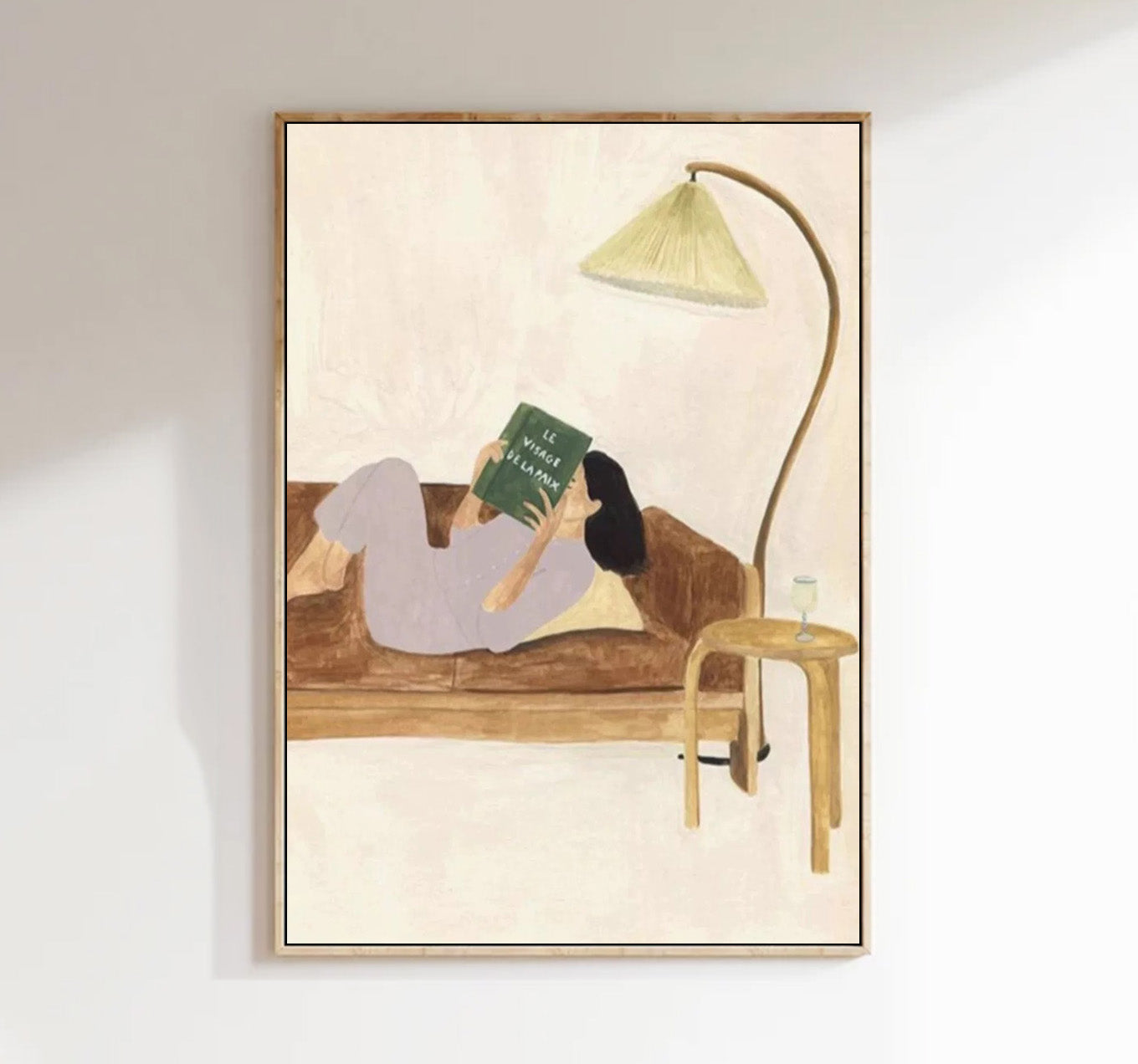 Framed artwork of a person reading a book on a couch with a lamp and glass.